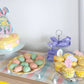 Pastel-colored Easter-themed dessert table with cake, cupcakes, and cookies.